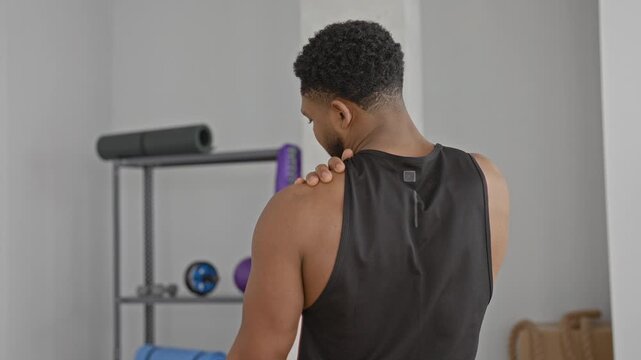 Man with hand on left shoulder after training, wearing black tank top in fitness studio with mat and rack of equipment; rehab calm.