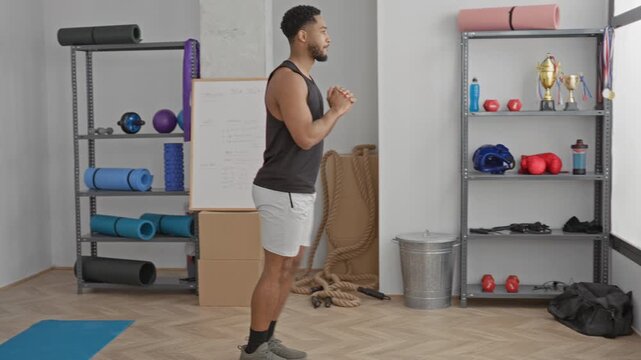 Man lunging with hands clasped, bare legs braced beside yoga mat and shelving with rope and trophy in studio; determination.