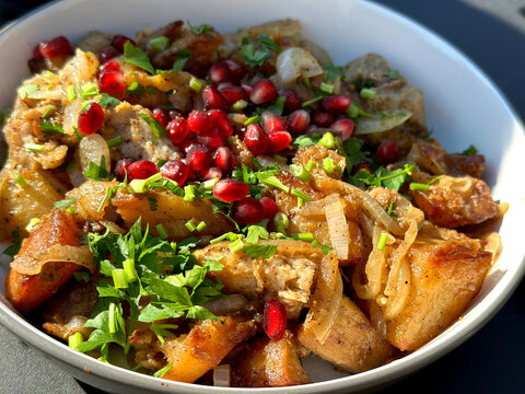 A close-up of a traditional dish featuring golden roasted potatoes, saut&eacute;ed onions, and meat, garnished with fresh parsley and vibrant red pomegranate seeds in a white bowl.