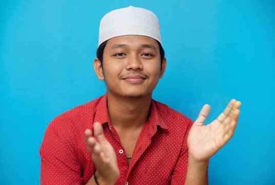 Young Muslim Man in White Skullcap Clapping Hands and Smiling on Blue Backdrop