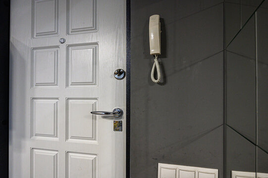 white six-panel entrance door with a chrome handle and lock; beside it, a beige wall-mounted intercom phone on a dark gray tiled wall with mirrored sections