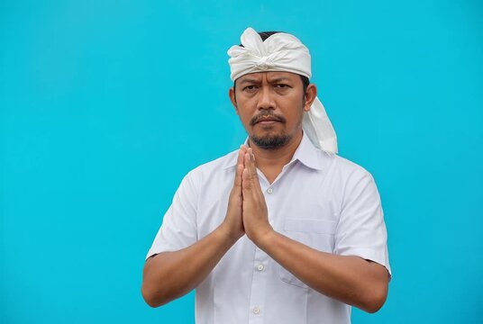 Traditional Balinese Man in White Attire Gesturing Respect on Blue Background