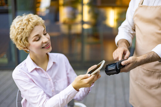 A woman smiles while making a contactless payment using her mobile phone at a point of sale terminal operated by a server