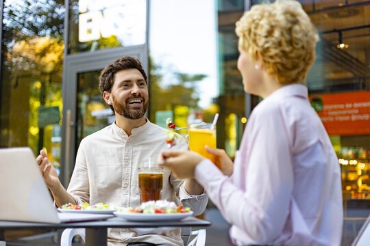 A man and woman are laughing and talking while having lunch and drinks at an outdoor cafe table with a laptop present