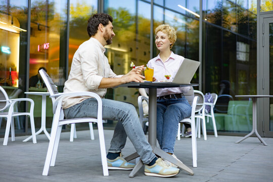 A man and woman are sitting at a table outside a modern cafe, enjoying drinks and conversation while a laptop is open