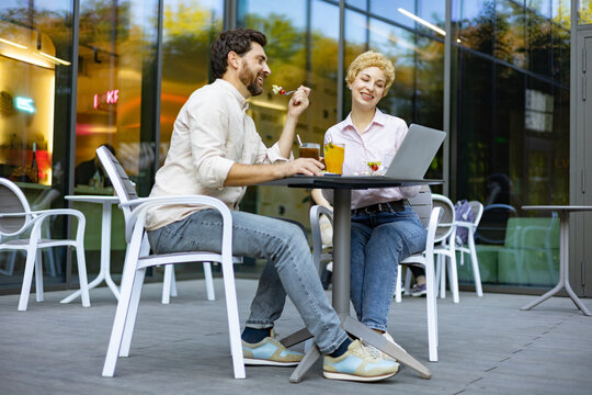 A man and woman are seated at a table outside a cafe, enjoying food and drinks while looking at a laptop