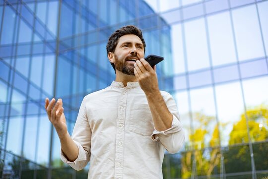 A man with a beard speaks into his smartphone held to his ear while gesturing with his other hand in front of a modern glass building