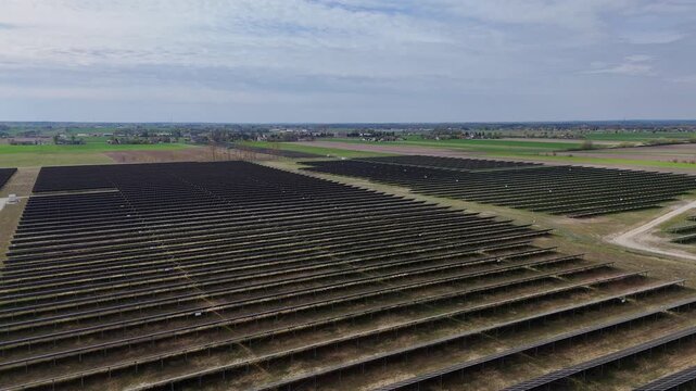 Aerial drone view of a vast solar farm featuring rows of photovoltaic panels arranged across an open rural field, overcast sky, renewable energy infrastructure, modern green technology landscape.