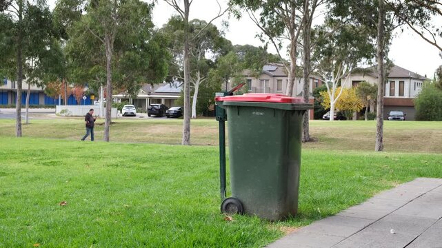 Australian council wheelie bin on a public green space beside a footpath in a suburban Melbourne neighbourhood.&nbsp;A pedestrian walks in the background.&nbsp;Waste collection infrastructure.