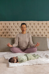 Obraz premium Mid-adult mother and infant daughter sitting meditating on bed with headboard, knit sweater, onesie