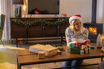 Naklejka premium Woman wearing Santa hat and patterned knit sweater wrapping kraft paper gifts in living room