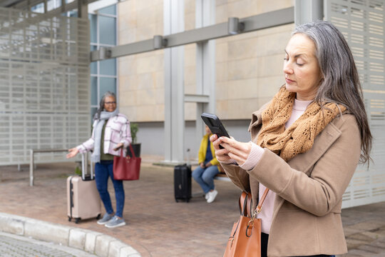 Mature woman tapping smartphone at curb, travel app map animating over suitcases guiding pickup