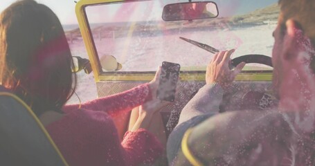 Naklejka premium Driving couple wearing pink sweater and gray jacket on sand in yellow buggy, holding smartphone