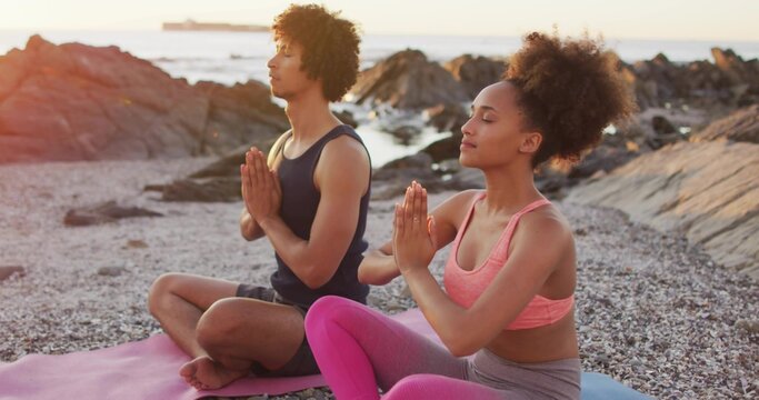 Meditating couple wearing sportswear sitting cross-legged on pebbly beach, pink and blue mats