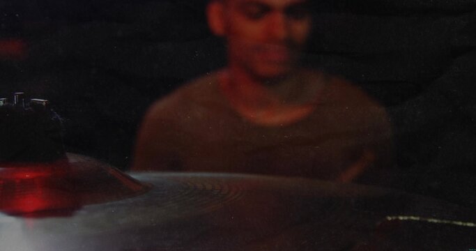Playing drummer wearing dark crewneck shirt sitting at drum kit in dim rehearsal, red-lit cymbal