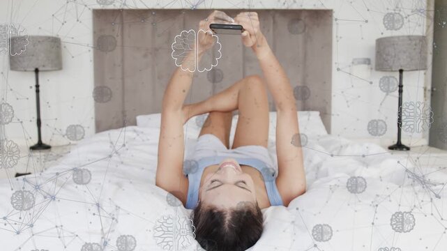 Woman reclining on bed lifting smartphone and swiping, viewing brain network visuals for tech demo