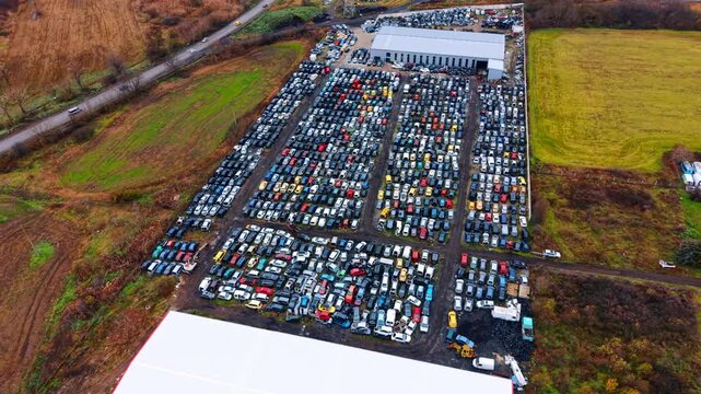 High angle aerial view of large automotive salvage yard. Wide shot of car junkyard with rows of vehicles next to industrial warehouse and rural road.
