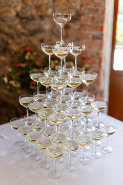 Pyramid of champagne coupes filled with sparkling wine on a white table, set against a warm stone wall and greenery
