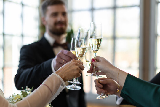 Bride, groom, and guests clink champagne flutes during an indoor wedding toast with soft window light and blurred background