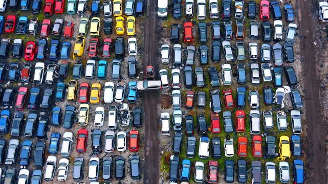 Industrial loader moving damaged vehicle in scrapyard. Drone shot showing a loader lifting a scrap car amidst rows of abandoned vehicles at a recycling yard.