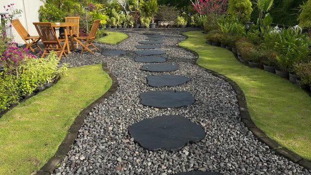 Curved path winds through manicured garden. Black stones contrast with gravel and green grass. Wooden chairs invite quiet relaxation. Lush plants line the path on both sides
