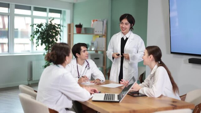 Positive young malaysian female medical student standing with papers in hands near table where sitting doctors and talking in medicals seminar in bright office 
