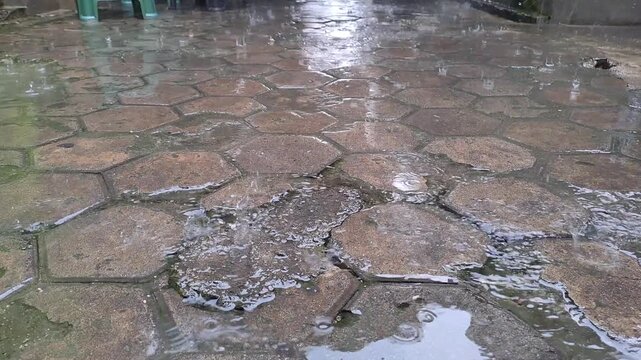 Handheld footage of tropical rain falling on hexagonal paving stones, wet street surface, natural rainfall pattern, moody rainy season atmosphere in a tropical country.