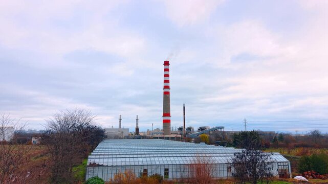 Aerial view of industrial greenhouse complex near power plant. Industrial greenhouse facility located next to a thermal power station with a red and white chimney under cloudy sky.