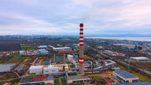 Industrial facility with tall chimney under overcast sky. Medium aerial view of a thermal power plant complex with a striped stack and industrial buildings.