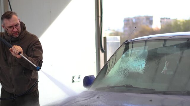 A middle aged man with eyeglasses washes a blue car at a self-service car wash. The car wash's high-pressure jet sprays water and rinses away the cleaning foam. 4k footage.