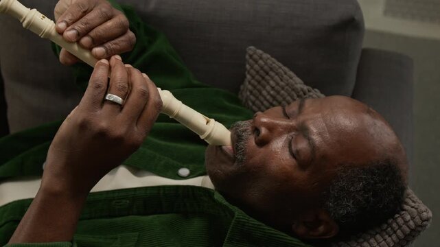 Close up shot of senior African American man with eyes closed playing recorder while lying on couch at home and enjoying music, copy space Great for music related service promotion