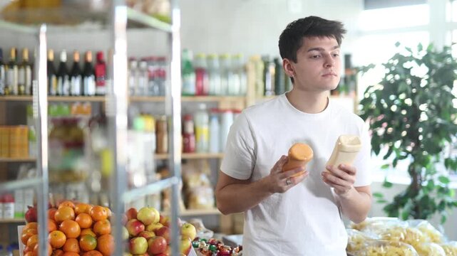 Positive thoughtful young guy reading labels on bottles with sauces in supermarket, carefully examining ingredients and expiration date while shopping for groceries