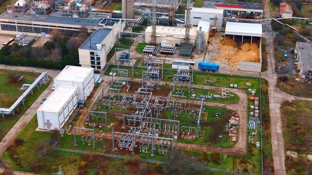 Top view of electrical substation with transformers and equipment. High angle shot of power distribution facility with metal structures, wires, and industrial buildings.