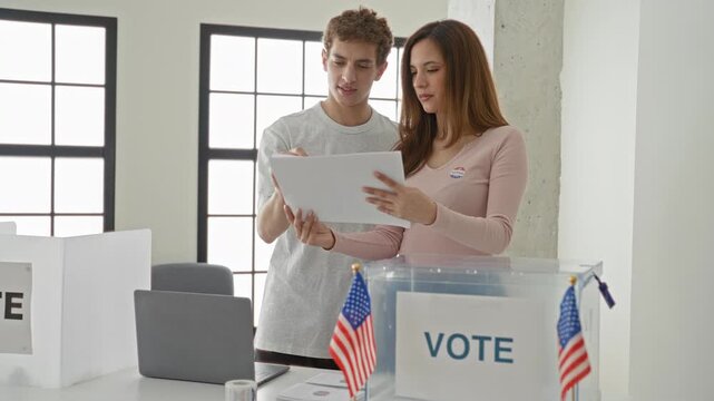 Woman holding ballot paper while man points to the paper beside a clear ballot box at a voting booth in a building; patriotic duty.