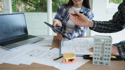 Architects reviewing house plans at desk with laptop, tablet and scale model, collaborative...