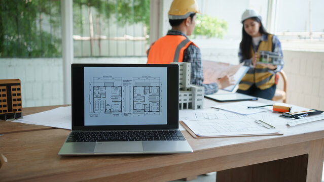 Architects or engineers in hard hats review blueprints at an office desk with laptop floor plan, scale model buildings, and documents, discussing construction project planning.