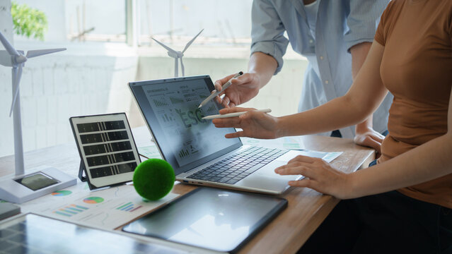 Two colleagues discuss ESG data on a laptop in a bright office with wind turbine models, tablet and reports, suggesting sustainable business, renewable energy planning and green finance.
