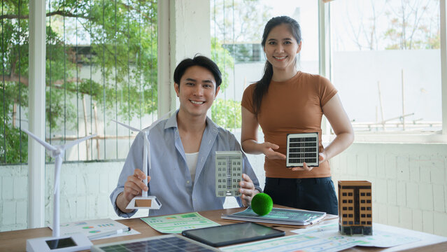 Two young professionals presenting solar panel models and building prototypes at a bright office desk, discussing renewable energy project planning, charts and documents on table