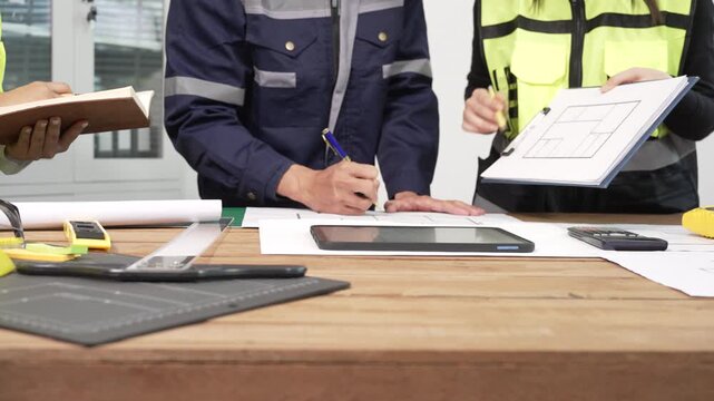 Construction team reviewing building floor plan at worktable with tools, tablet and documents; engineers and supervisors discussing project details in bright office.
