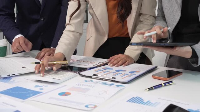 Business team meeting at office desk analyzing charts and financial reports, hands using calculator and tablet for planning and strategy.