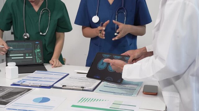Healthcare team meeting at desk reviewing medical charts and tablet X-ray, with doctors in scrubs and lab coat discussing patient data, analytics and planning treatment.
