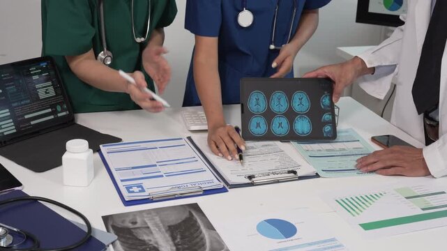 Medical team meeting at desk reviewing digital tablet with brain scan images, charts and documents; doctors and nurses in scrubs and lab coat discussing patient data, modern healthcare collaboration