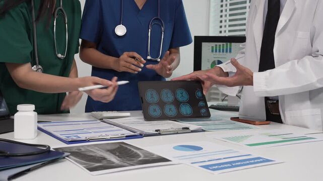Medical team meeting at desk reviewing digital tablet with brain scan images, charts and documents; doctors and nurses in scrubs and lab coat discussing patient data, modern healthcare collaboration