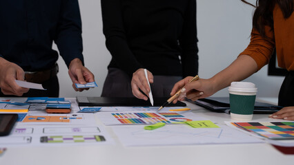 Fototapeta premium Design team collaborating over desk with printed charts, color palettes, sticky notes and coffee cup, hands pointing and writing in office meeting.