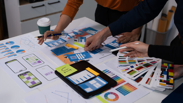 Team of coworkers reviewing printed design mockups and charts on a table in an office, collaborating on branding or marketing strategy with coffee cups and stationery.