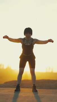 Tattooed woman performing lateral raises with dumbbells at sunset