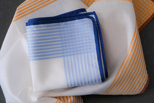 Close-up folded white handkerchief with blue stripes and hand-rolled edge on unfolded orange striped border handkerchief.