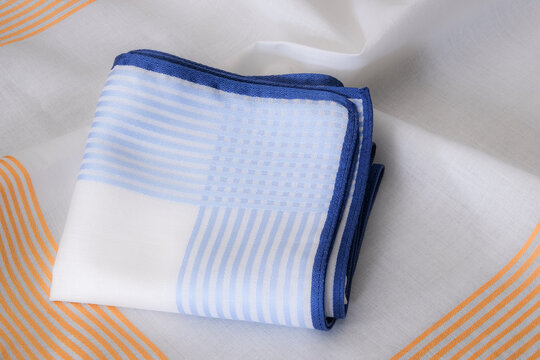 Close-up folded white handkerchief with blue stripes and hand-rolled edge on unfolded orange striped border handkerchief.