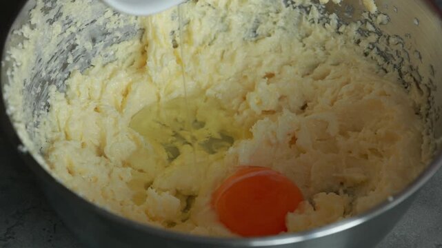 Close-up of a child's hands adding an egg into a butter and sugar mixture while using a hand mixer