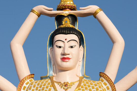 Close-up of a multi-armed Buddhist deity statue with an ornate golden headdress and serene expression against a bright blue sky, highlighting intricate details and spiritual tranquility.